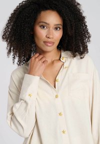 Crème-kleurige langwerpige blouse met lange mouwen, voorzien van gele sterrenversieringen op de knopen en een borstzak. Zachte, gestructureerde stof.