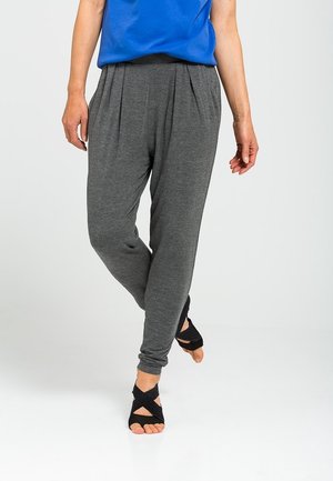 Persoon draagt grijze losse broek en zwarte open teen dansschoenen, staat met één been gekruist over het andere.