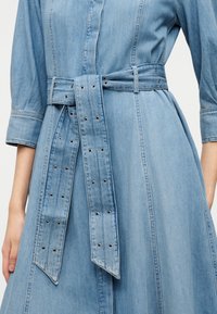 Φόρεμα από τζιν σε ανοιχτό γαλάζιο χρώμα, με σφηκοφωλιά στη μέση και ζώνη που ταιριάζει. Η ζώνη διαθέτει μεταλλικές οπές και μυτερές άκρες. Μακριά μανίκια και κουμπιά στο μπροστινό μέρος.