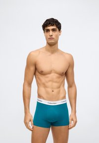 Muž bez trička s krátkymi kučeravými tmavými vlasmi oblečený v tyrkysových boxerkách Calvin Klein s bielym logom na páske, stojaci na pozadí s plnou bielou farbou.