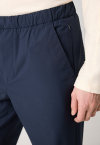 Navy blauwe broek met elastische tailleband en zijzakken. Glad, lichtgewicht stof met een lichte glans en een getailleerde pasvorm. Close-up van het heupgebied.
