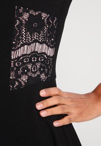 Handen vilar på höften av en svart klänning med genomskinlig svart spetsblompanel över blekrosa tyg.