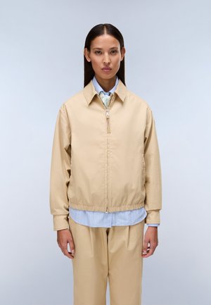 Kobieta o długich ciemnych włosach, ubrana w beżową kurtkę na suwak, na jasnoniebieską koszulę i pasujące beżowe spodnie, stoi na tle jednolitego jasnego tła.