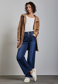 Beżowa kurtka zapinana na zamek w połączeniu z białą bluzką, zestawiona z niebieskimi spodniami o szerokich nogawkach i białymi trampkami. Kurtka ma kaptur i luźny krój.