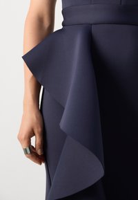 Robe de marine avec un ourlet à volants superposés, une taille structurée et un tissu lisse. Main visible avec une bague en argent reposant sur le côté.
