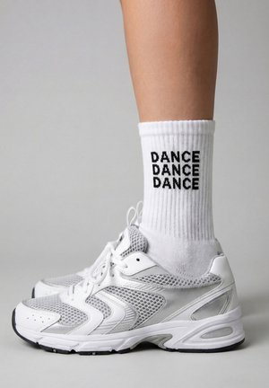Jambe inférieure portant une chaussure de sport blanche et une chaussette côtelée blanche avec le mot « DANCE » répété trois fois en texte noir.
