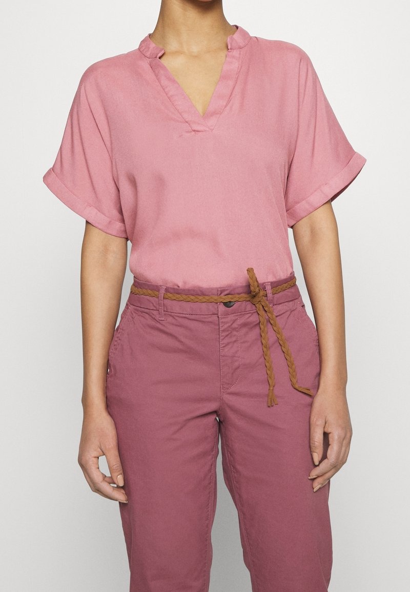 Personne portant un chemisier rose clair à manches courtes et col en V, rentré dans un pantalon taille haute mauve avec une ceinture tressée marron nouée devant.