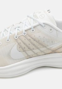 Chaussure de course légère avec une tige en maille beige et blanche, dotée d'une semelle lisse, d'accents gris et d'un design à motif texturé.