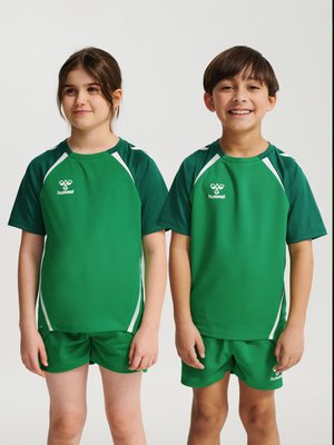 Twee kinderen staan zij aan zij, gekleed in bijpassende groene sportshirts en shorts, glimlachend tegen een effen achtergrond.