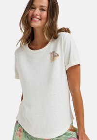 Camiseta blanca de manga corta con un cuello redondeado y una textura ligera. Presenta un diseño floral bordado en tonos dorados y marrones.