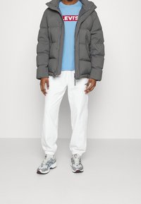 Szara kurtka puchowa, jasnoniebieski t-shirt z czerwonym logo, białe spodnie oraz srebrne trampki z niebieskimi akcentami i czerwonymi detalami.