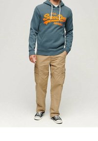 Hombre vistiendo un hoodie teal de SuperDry, pantalones cargo color beige y zapatillas negras, de pie con una mano en el bolsillo, frente a un fondo liso.