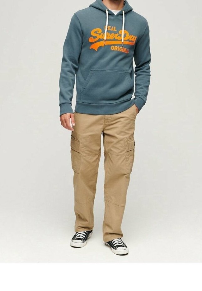 Hombre vistiendo un hoodie teal de SuperDry, pantalones cargo color beige y zapatillas negras, de pie con una mano en el bolsillo, frente a un fondo liso.