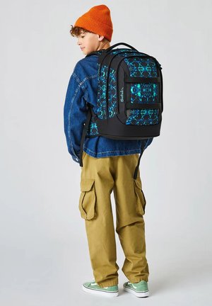 Ragazzo che indossa un berretto arancione, giacca di jeans, pantaloni cargo kaki e scarpe da ginnastica verdi, con uno zaino grande nero e blu a motivo.