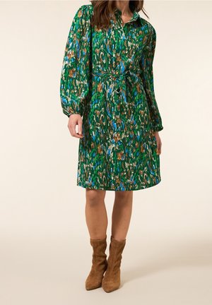 Mujer vistiendo un vestido de manga larga con estampado en verde, azul y marrón, con cinturón en la cintura, combinado con botines de ante marrones.