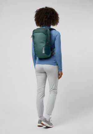 Grüner Rucksack mit schlankem Design, gepolsterter Rückseite, verstellbaren Trägern und Reißverschlusstaschen. Getragen über einem blauen Langarmoberteil.