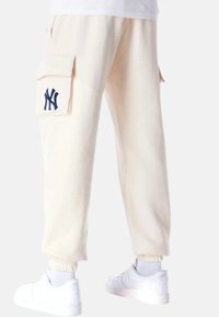 Lys beige cargo bukser med store sidelommer, et marineblåt broderet logo og elastiske manchetter. Kombinationen er med hvide sneakers.