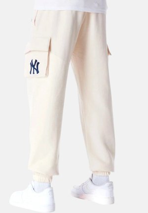 Pantalones cargo de color beige claro con grandes bolsillos laterales, un logo bordado en azul marino y puños elásticos. Combinados con zapatillas blancas.