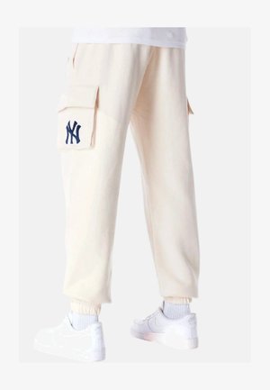 Helle beige Cargo-Hosen mit großen Seitentaschen, einem navyblauen gestickten Logo und elastischen Bündchen. Kombiniert mit weißen Sneakern.