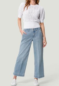 Suéter corto de punto blanco con un patrón texturizado, combinado con jeans de pierna ancha azul claro. La modelo lleva zapatillas blancas.