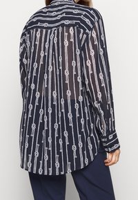 Camicia blu navy con motivo a corda bianca, tessuto leggero e trasparente, maniche lunghe, polsini con bottoni e colletto. Il retro presenta un design a strisce verticali.