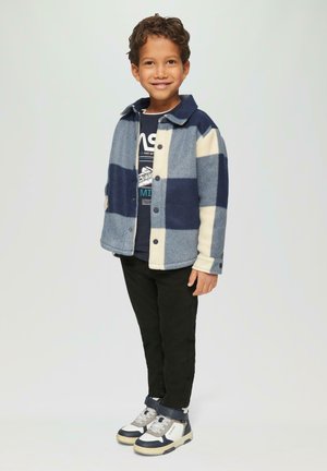 Veste en laine à carreaux bleus et crème, assortie à un t-shirt graphique bleu marine, un pantalon noir et des baskets montantes grises et bleu marine.