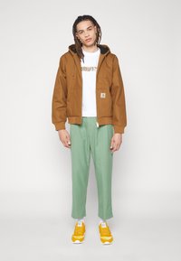 Brown hooded jacket with front zipper, white t-shirt featuring a logo, green trousers, and yellow sneakers with white accents.