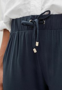 Pantalone u tamnoplavoj boji s elastičnim pojasom i naboranom teksturom, s vezicom i zlatnim metalnim vrhom na vezici.