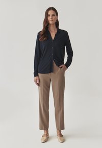 Navy-blauwe blouse met lange mouwen, gecombineerd met beige rechte broek en lichte schoenen, staande tegen een neutrale achtergrond.