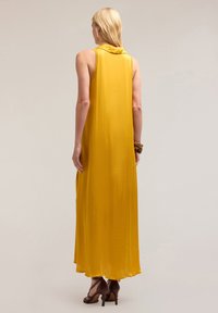Rochie maxi galbenă fără mâneci, cu un design drapat la spate, realizată dintr-un material neted, având guler rotund și o siluetă fluidă.