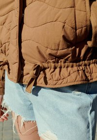 Bruine gewatteerde jas met een trekkoord in de taille, gecombineerd met lichtblauwe gescheurde denimshorts. Textuurstof met zichtbare stikseldetails.