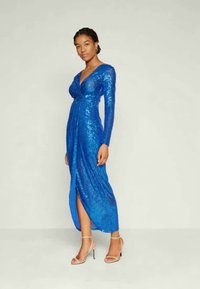 Μακριά μπλε φόρεμα με παγιέτες που διαθέτει V-λαιμόκοψη, μακριά μανίκια, εφαρμοστή γραμμή σώματος και σκίσιμο ψηλά στο μηρό. Συνδυασμένο με σανδάλια με ψηλά τακούνια και λουράκια.