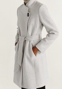 Manteau gris clair en tissu texturé, avec un col montant, un grand bouton unique et une ceinture à nouer à la taille ; doté de poches latérales.