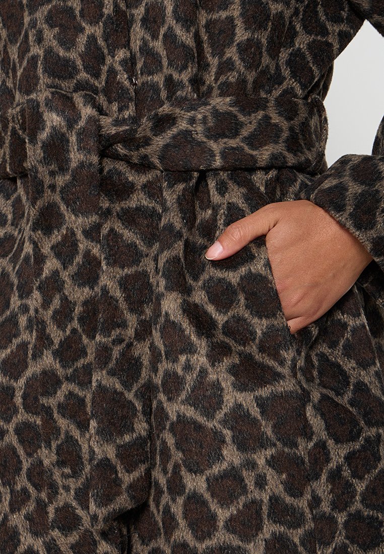 Mano che riposa nella tasca di un cappotto cinturato maculato marrone e nero con tessuto strutturato.