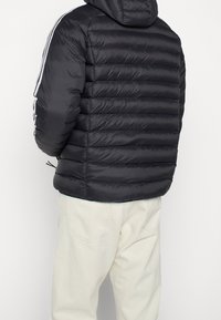 Svart puffjacka med horisontell sömnad, huva och vita sidostråk. Har elastiska ärmslut och justerbar nederkant för en säker passform.