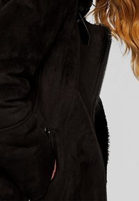 Cappotto in finta pelliccia nera con una texture liscia, dotato di chiusura con zip e tasche laterali, che mette in mostra un design aderente e una morbidezza unica.