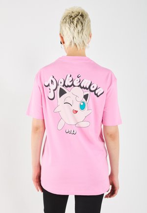 Osoba z krótkimi blond włosami ubrana w różowy t-shirt z puszczającym oczko Jigglypuffem i słowem „Pokémon” na plecach.