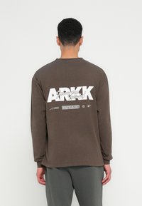 Brahk dolgimi rokavi majica s krožnim izrezom, ki ima na sprednji strani velike bele napise "ARKK" in manjši napis na hrbtni strani. Iz bombažnega blaga, sproščen kroj.