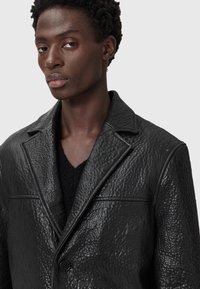 Schwarze strukturierte Lederblazer mit einem Knopfverschluss, Notchkragen und dezenten Ziernähten. Getragen über einem schwarzen Pullover.