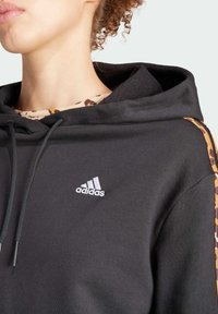 Černá mikina s kapucí z texturované látky, která má šedou tkaničku a reliéfní bílé logo. Boční panely vykazují vzor leopardího tisku.