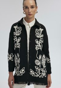 Cardigan nero con ricami floreali bianchi, chiusura con bottoni, colletto e orlo frangiato. Presenta maniche lunghe e un tessuto testurizzato.
