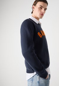 Sweatshirt azul marinho feito de algodão, com bordado laranja "BAND" no peito. Punhos e bainha canelados, usado por cima de uma camisa branca com colarinho.