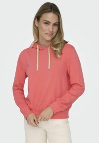 Sudadera color coral con cordón y detalles dorados. Presenta un pequeño corazón dorado en el pecho, hecha de tela suave con un corte relajado.