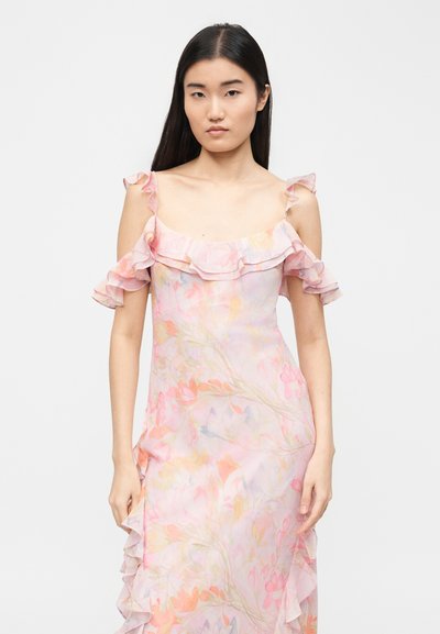 Νεαρή γυναίκα με μακριά μαύρα μαλλιά, ντυμένη με ροζ παστέλ φόρεμα με φλοράλ σχέδιο, με αποκαλυπτικούς ώμους και βολάν στα μανίκια και στη λαιμόκοψη.