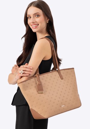 Junge Frau mit langen braunen Haaren lächelt, trägt eine große beige gemusterte Tragetasche über der Schulter und ist in einem ärmellosen schwarzen Oberteil gekleidet.