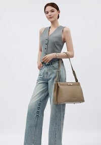 Beige strukturierte Handtasche mit Textur, goldenen Hardware-Akzenten, Handtragriemen und einem Crossbody-Tragegurt, präsentiert neben hellblauen weiten Jeans und einem grauen ärmellosen Top.