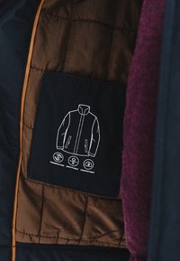 Wyściełana, granatowa kurtka z brązowym, pikowanym wnętrzem oraz graficzną metką na kieszeni, która przedstawia zarys kurtki z trzema ikonami poniżej.