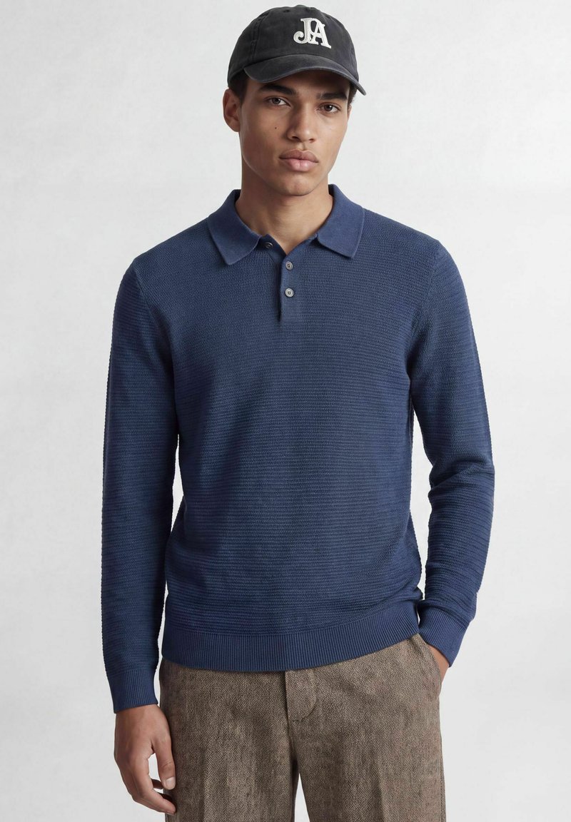 Jeune homme portant un polo bleu foncé à manches longues, un pantalon marron et une casquette noire avec des lettres blanches JA.