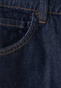 Tissu en denim bleu foncé avec des coutures orange visibles autour d'une poche avant et d'une passants de ceinture.