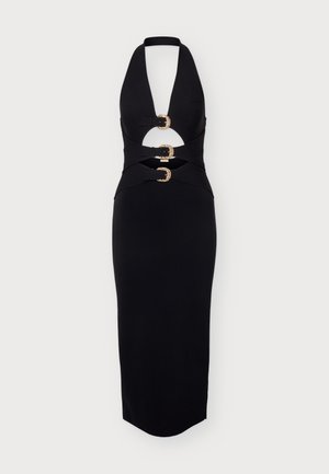 Un mannequin porte une robe midi noire sans manches, ornée de trois boucles dorées sur le devant, ainsi que des escarpins noirs à bout pointu. Il se tient devant un fond clair uni.